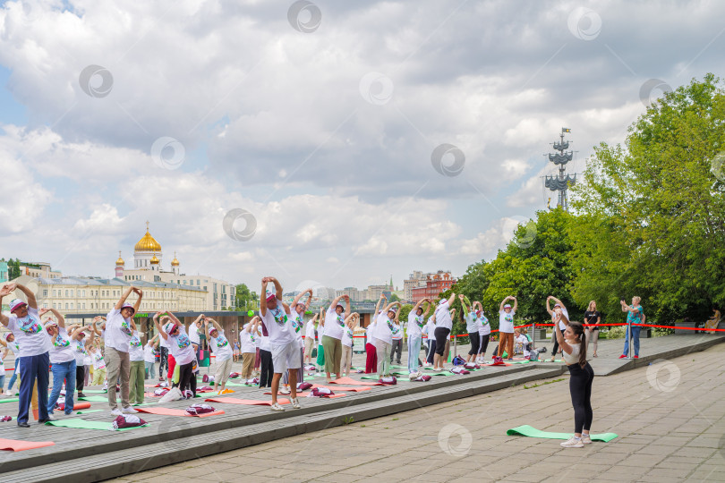 Скачать Пенсионеры, участники спортивного праздника программы "Московское долголетие" в фирменных московских футболках и шапочках выполняют гимнастику под руководством опытного тренера в парке Музеон на фоне храма Христа Спасителя, голубого неба и кучев... фотосток Ozero