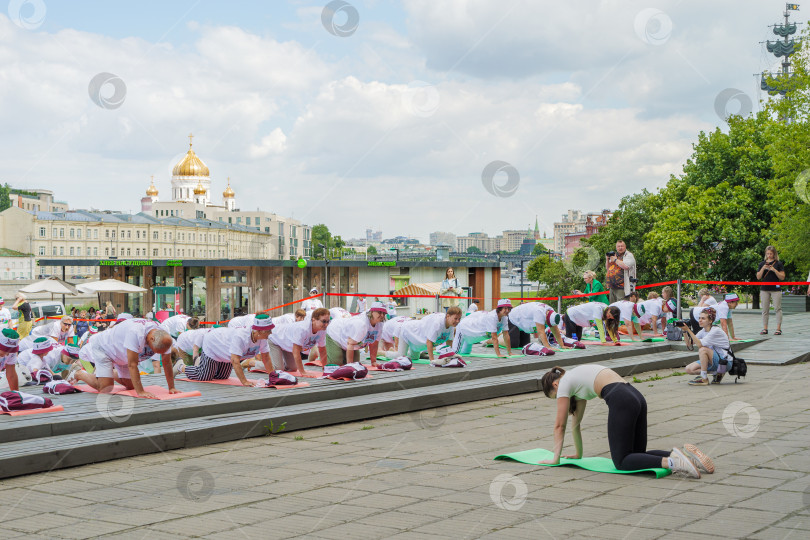 Скачать Пенсионеры, участники спортивного праздника программы "Московское долголетие", в фирменных московских футболках и шапочках, опустившись на разноцветные гимнастические коврики, выполняют гимнастику под руководством тренера в парке Музеон на фоне... фотосток Ozero