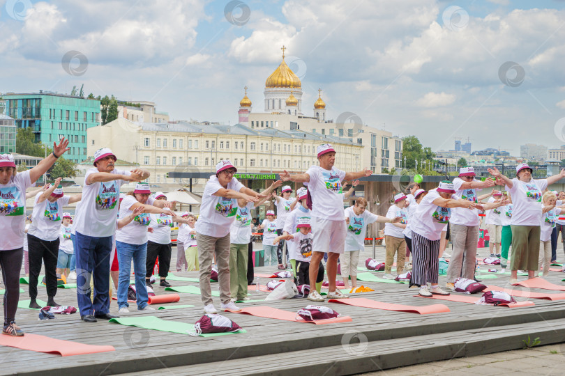 Скачать Мужчины и женщины пенсионного возраста в фирменных московских футболках и головных уборах в московском парке Музеон выполнят гимнастику, стоя на разноцветных гимнастических ковриках на фоне храма Христа Спасителя, голубого неба и кучевых облаков... фотосток Ozero