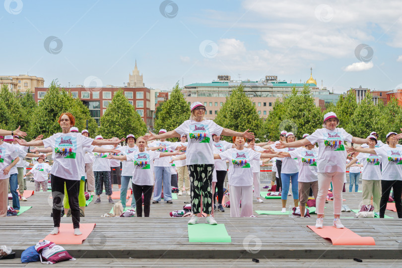 Скачать Женщины пенсионерки в фирменных московских футболках и головных уборах в московском парке Музеон выполнят гимнастику, стоя на разноцветных гимнастических ковриках. фотосток Ozero