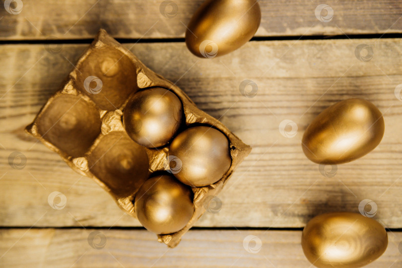 Скачать Вид сверху Пасхальные яйца, раскрашенные золотой краской, в бумажном контейнере на деревянном фоне. Праздничные украшения. фотосток Ozero