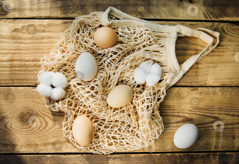 Скачать Концепция Пасхи. Бежевая сетчатая хозяйственная сумка с коричневыми куриными яйцами на деревянном фоне. фотосток Ozero