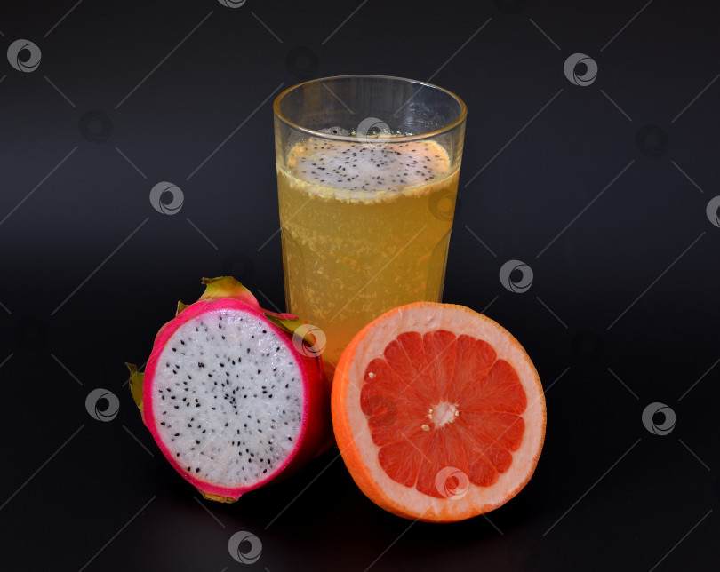 Скачать Свежевыжатый сок из тропических фруктов с косточками в высоком граненом стакане на черном фоне, рядом с половинками спелой питайи и грейпфрута. фотосток Ozero