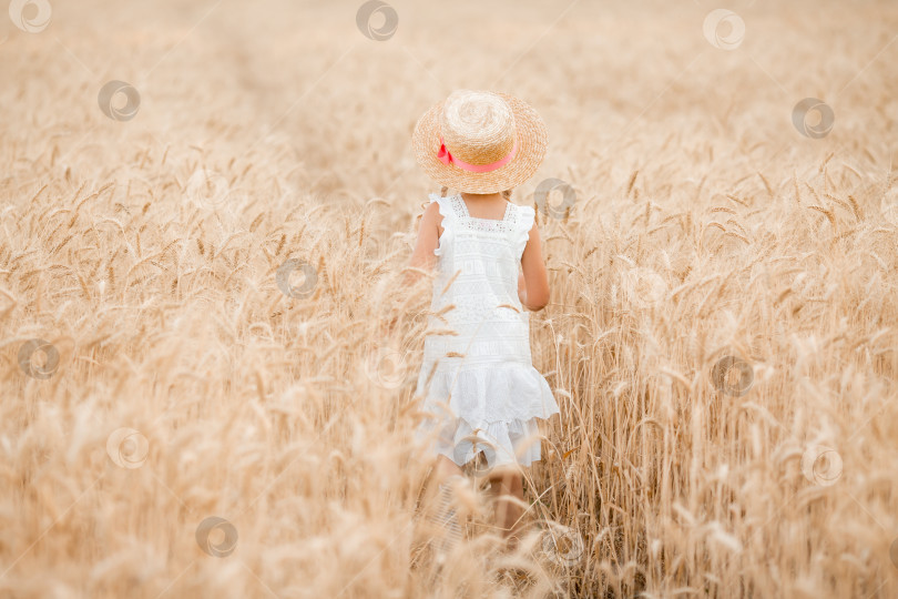 Скачать Маленькая девочка в соломенной шляпке и хлопчатобумажном платье с пучком бежит по пшеничному полю летом фотосток Ozero