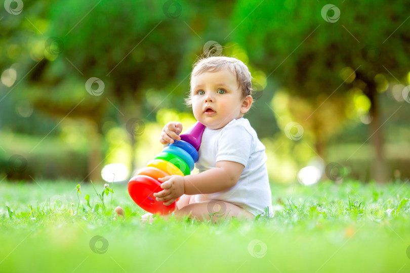 Скачать Симпатичный малыш-мальчик в белом боди сидит летом на зеленой траве и играет с платьем разноцветной пирамидки. Ребенок играет с развивающей игрушкой. Расставание с ребенком в возрасте до года фотосток Ozero