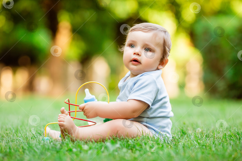 Скачать Симпатичный маленький мальчик в синем боди сидит летом на траве и пьет воду из бутылки. Здоровый малыш 10 месяцев сидит на траве и пьет воду из бутылочки фотосток Ozero