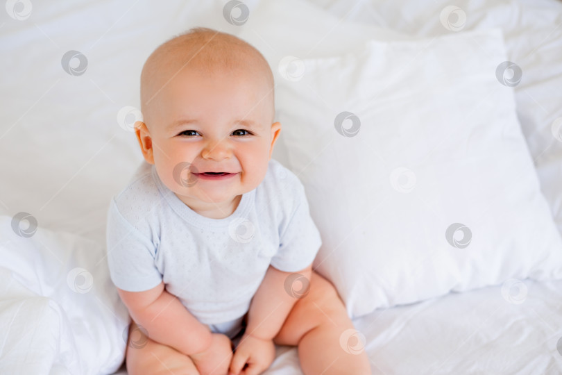 Скачать Милый мальчик 6 месяцев улыбается в белом боди, сидя на кровати на белом постельном белье фотосток Ozero