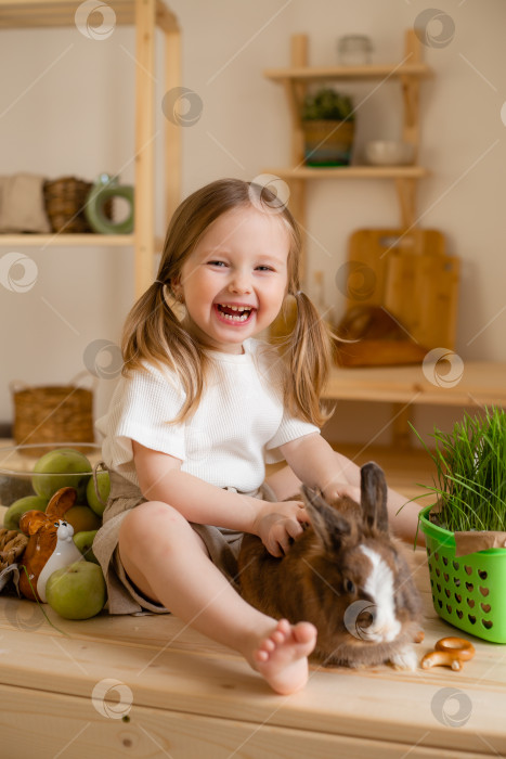 Скачать Милая маленькая девочка на деревянной кухне дома кормит кролика свежей травой фотосток Ozero