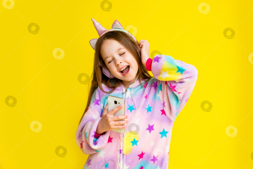 Скачать счастливая маленькая девочка в кигуруми с единорогом лежит на желтом фоне среди разноцветных конфетти и слушает музыку фотосток Ozero