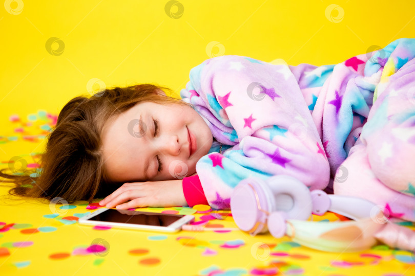 Скачать счастливая маленькая девочка в кигуруми с единорогом лежит на желтом фоне среди разноцветных конфетти и слушает музыку фотосток Ozero