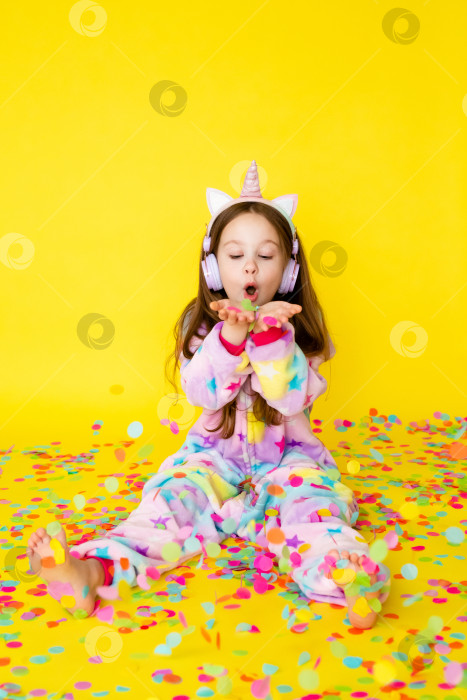 Скачать маленькая девочка с длинными волосами, одетая в кигуруми и наушники в форме единорога, с удовольствием ловит конфетти, стоя на желтом фоне фотосток Ozero
