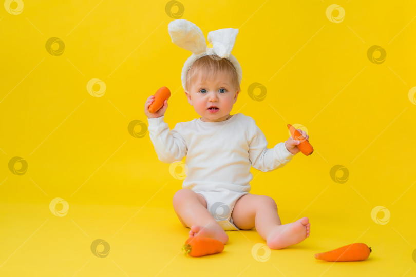 Скачать Малыш в белом боди с кроличьими ушками на голове ест морковь, сидит на желтом фоне с овощами. малышка в виде пасхального кролика, место для текста фотосток Ozero