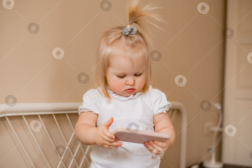 Скачать Малышка блондинка играет с телефоном дома на кровати. Дети и технологии фотосток Ozero