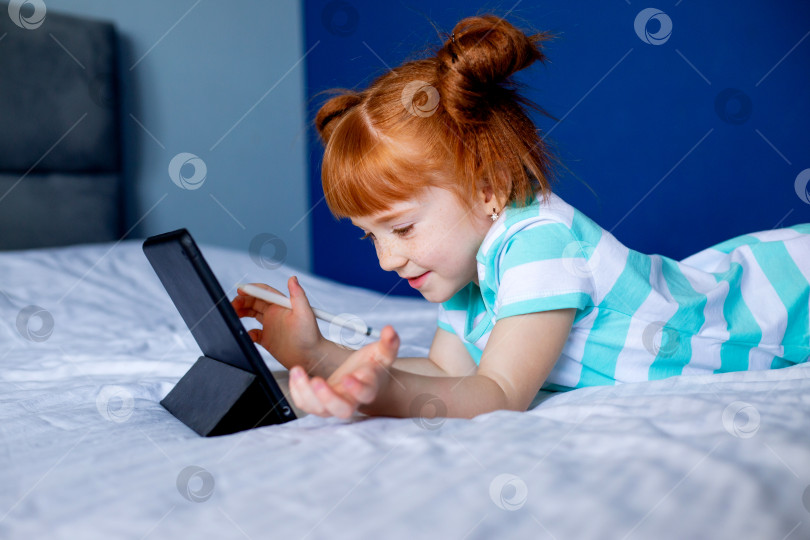 Скачать детский портрет девочки с рыжими волосами, лежащей на кровати с планшетом. дистанционное образование для детей фотосток Ozero