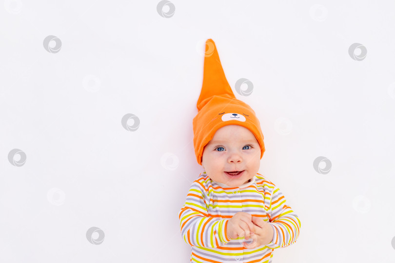 Скачать маленькая улыбающаяся девочка 6 месяцев, лежащая на белом изолированном фоне в оранжевой шапочке-колпачке, место для текста фотосток Ozero