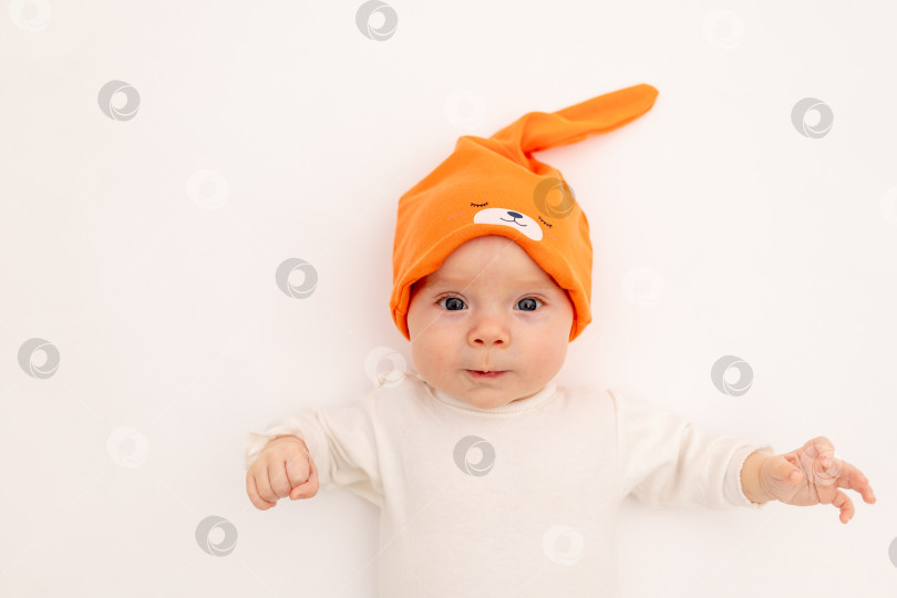 Скачать улыбающаяся малышка в белом боди и ярко-оранжевой шапочке лежит на белом изолированном фоне, портрет ребенка 3-6 месяцев, место для текста. Здоровый счастливый ребенок. фотосток Ozero