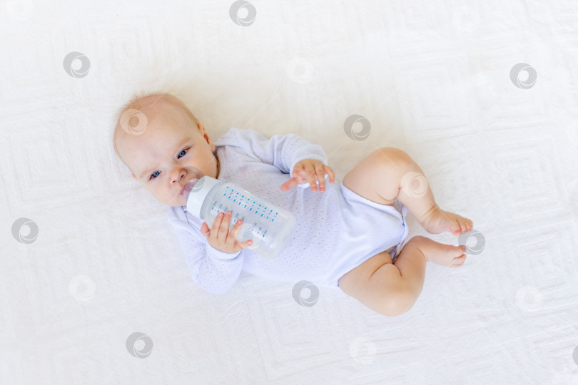 Скачать маленькая девочка 6 месяцев в белом боди лежит на спине на белой кровати дома с бутылочкой воды, концепция детского питания фотосток Ozero