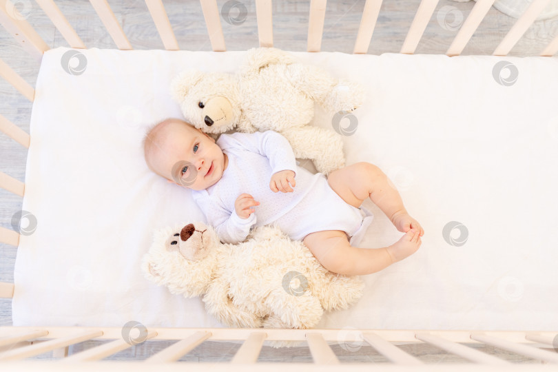 Скачать маленькая девочка 6 месяцев от роду в белом боди лежит на спине в детской кроватке с мягкими игрушками мишками фотосток Ozero