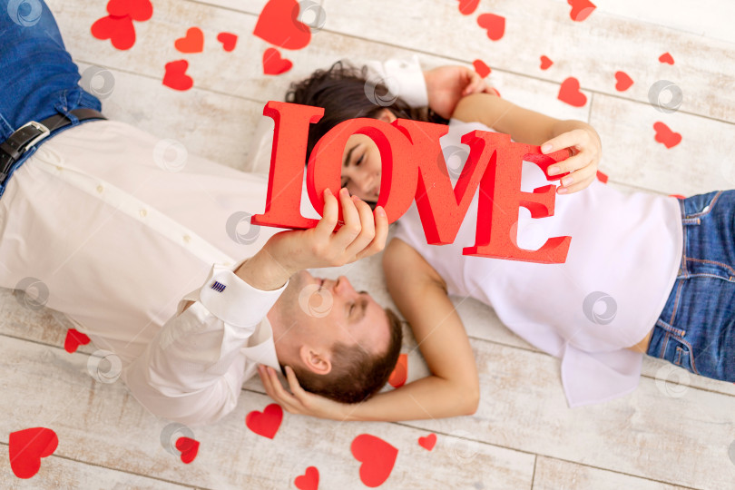 Скачать день святого Валентина, влюбленная пара лежит среди сердец на полу и держит большую надпись Love фотосток Ozero