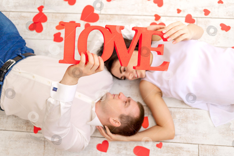 Скачать день святого Валентина, влюбленная пара лежит среди сердец на полу и держит большую надпись Love фотосток Ozero