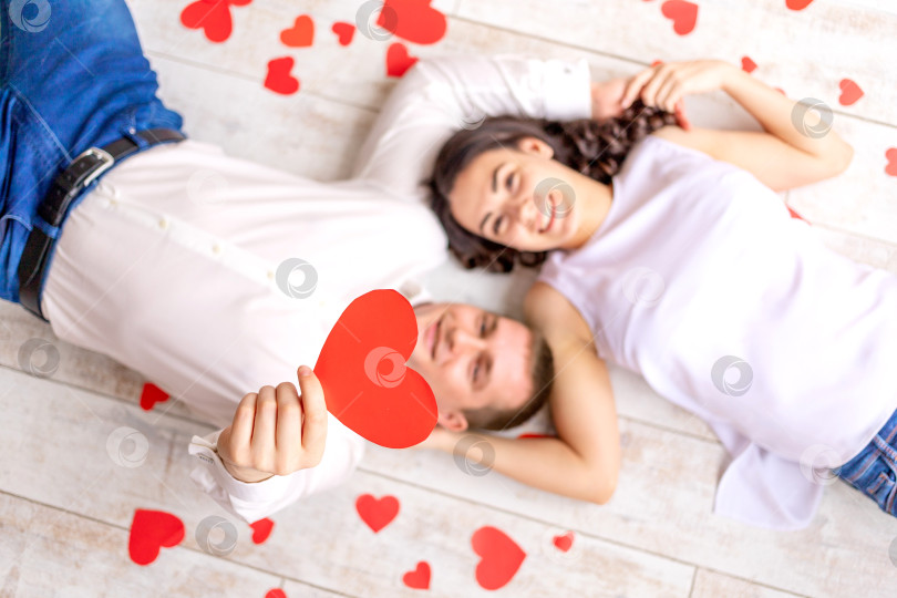 Скачать день святого Валентина, влюбленная пара лежит среди сердец на полу, обнимаясь и держа в руках большое красное сердце фотосток Ozero