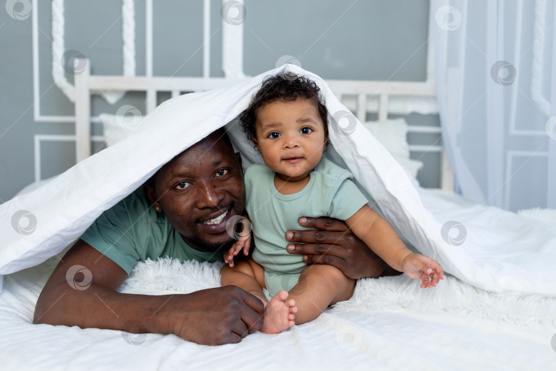 Скачать счастливый улыбающийся афроамериканский папа с маленьким сыном дома на кровати, обнимающиеся под одеялом, счастливая семья, день отца фотосток Ozero