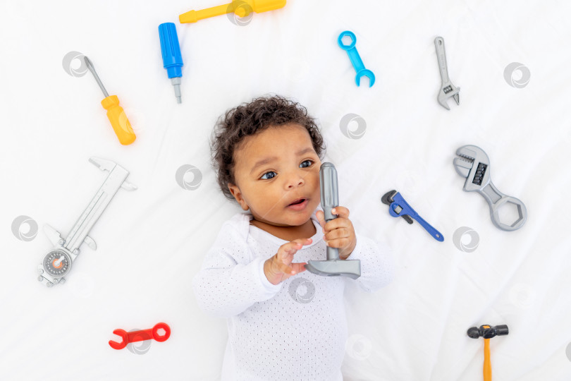 Скачать Афроамериканский маленький ребенок со строительными инструментами, лежащий на белой кровати, концепция строительства и ремонта фотосток Ozero