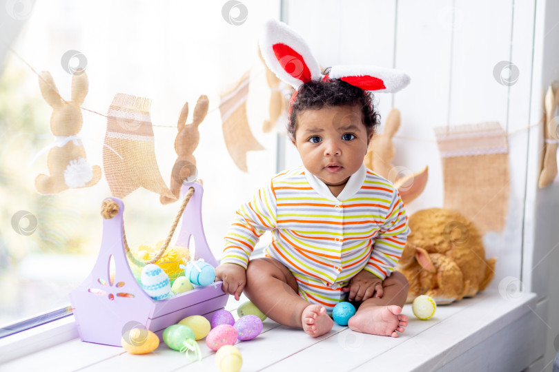 Скачать Концепция Пасхи, афроамериканский маленький ребенок в ушках зайца, на голове яйца и кролик на окне дома фотосток Ozero