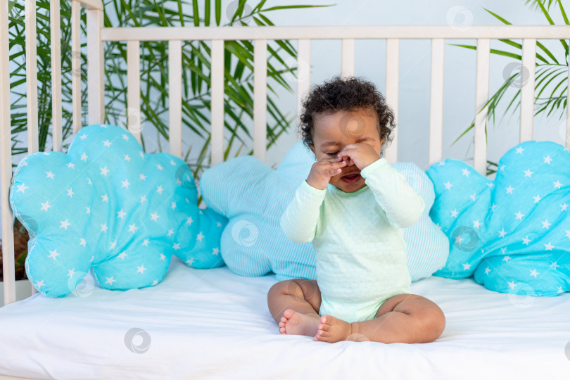 Скачать африканский младенец трет глаза, сидя дома в кроватке перед отходом ко сну, и хочет спать или плачет, закрыв лицо руками - концепция здорового сна фотосток Ozero