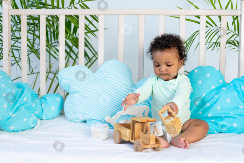 Скачать симпатичный африканский малыш сидит дома в кроватке на голубой хлопчатобумажной кровати и играет с деревянными игрушками фотосток Ozero
