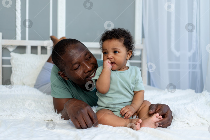 Скачать афроамериканский папа играет в погремушку с маленьким сыном дома на кровати в спальне, отцовская любовь фотосток Ozero
