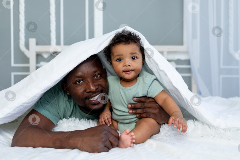 Скачать счастливый улыбающийся афроамериканский папа с маленьким сыном дома на кровати, обнимающиеся под одеялом, счастливая семья, день отца фотосток Ozero