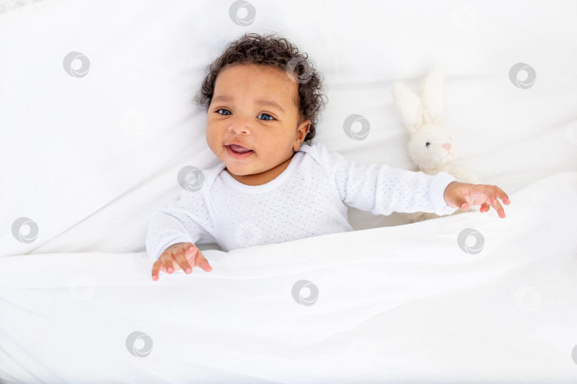 Скачать Улыбающийся маленький ребенок-афроамериканец под одеялом с игрушкой на белой кровати для сна фотосток Ozero