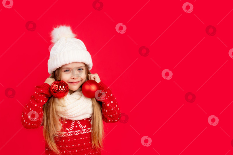 Скачать девочка-ребенок в зимней шапке и свитере с елочными шариками в качестве сережек в ушах на красном монохромном изолированном фоне радуется и улыбается, концепция нового года и Рождества, место для текста фотосток Ozero
