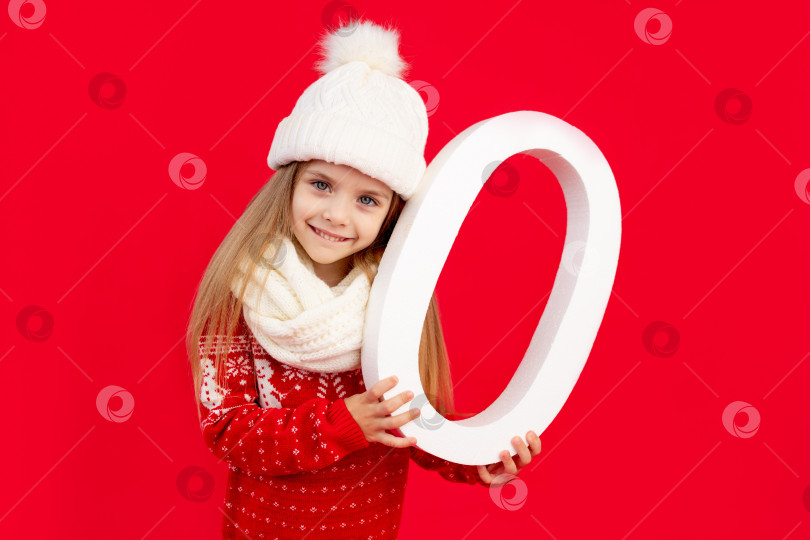 Скачать девочка-ребенок в зимней шапке и свитере с большой цифрой ноль на красном монохромном изолированном фоне радуется и улыбается, концепция нового года и Рождества, место для текста фотосток Ozero