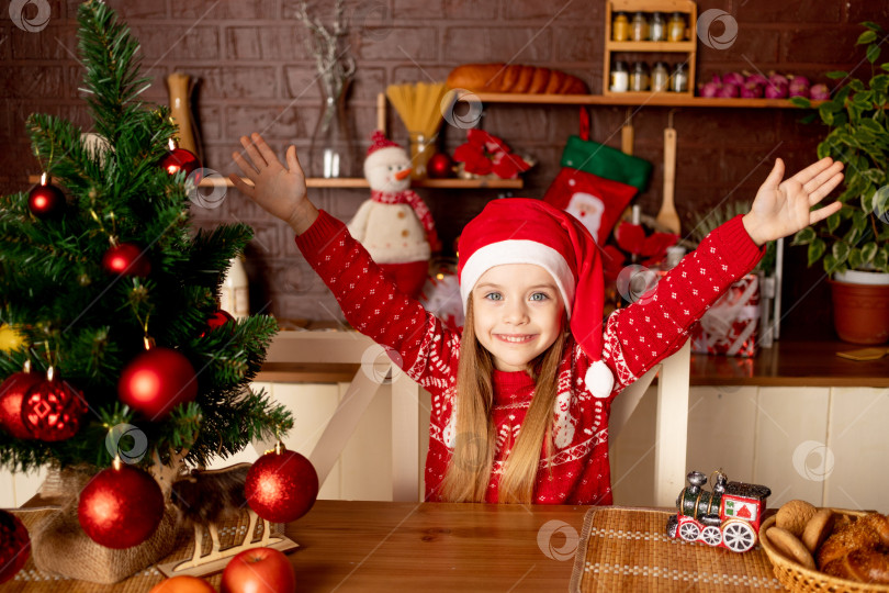 Скачать счастливая девочка-ребенок в шапочке Санты на темной кухне у рождественской елки с красными шарами радуется и улыбается - концепция нового года и Рождества фотосток Ozero