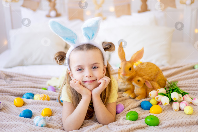 Скачать пасха, маленькая девочка среди раскрашенных яиц в заячьих ушках готовится к празднику, улыбаясь и веселясь фотосток Ozero