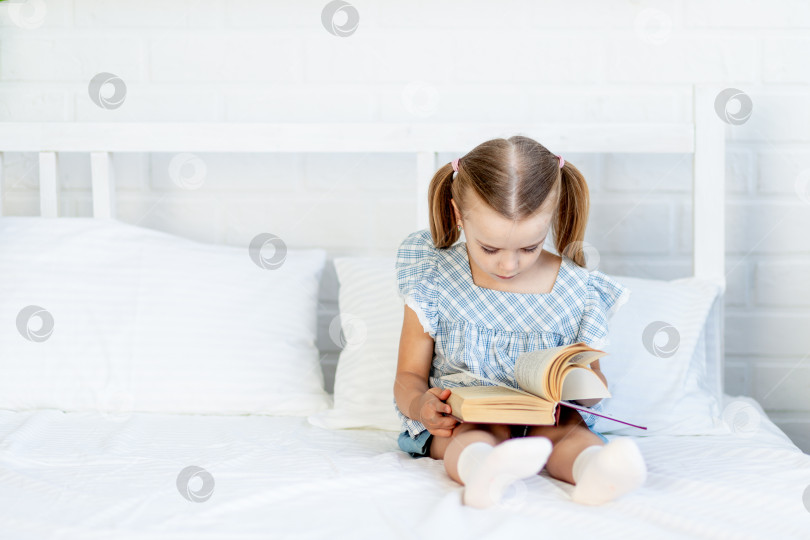 Скачать девочка читает книгу на кровати дома, сидя на белой хлопчатобумажной кровати. фотосток Ozero