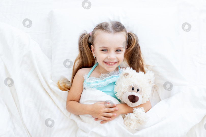Скачать маленькая девочка спит или проснулась утром дома на белой хлопчатобумажной кровати, обнимая в руках плюшевого мишку и мило улыбаясь фотосток Ozero