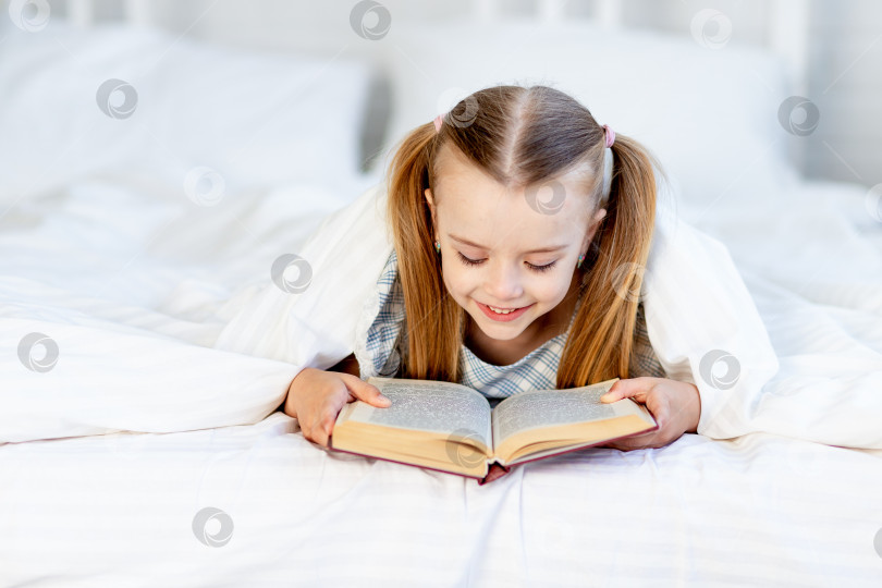 Скачать девочка читает книгу дома на белой хлопчатобумажной кровати под одеялом и мило улыбается фотосток Ozero