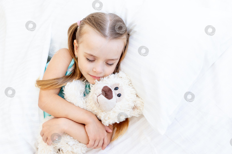Скачать девочка спит дома на кровати из белого хлопка, сжимая в руках плюшевого мишку и сладко улыбаясь во сне фотосток Ozero
