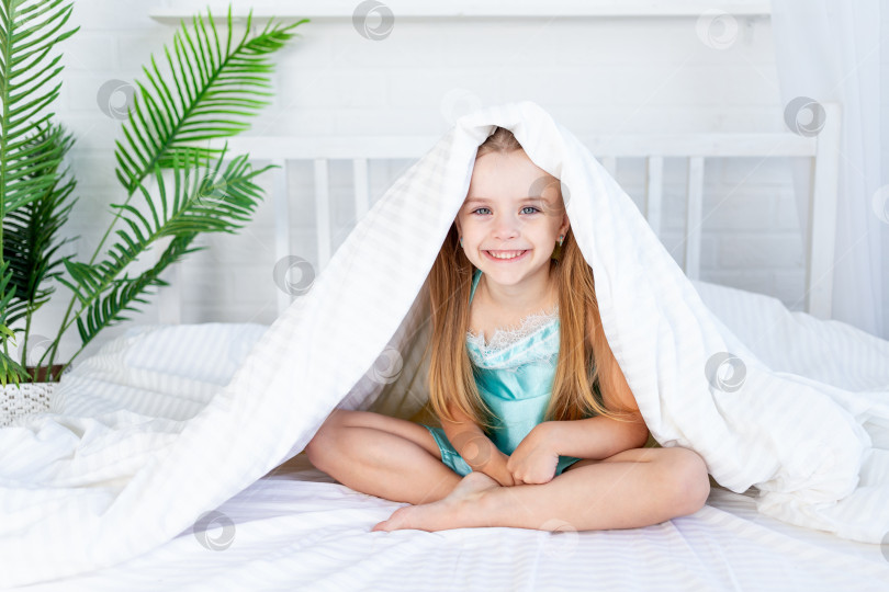 Скачать маленькая девочка играет под одеялом, просыпаясь утром дома на кровати, застеленной белым хлопком, и мило улыбается фотосток Ozero