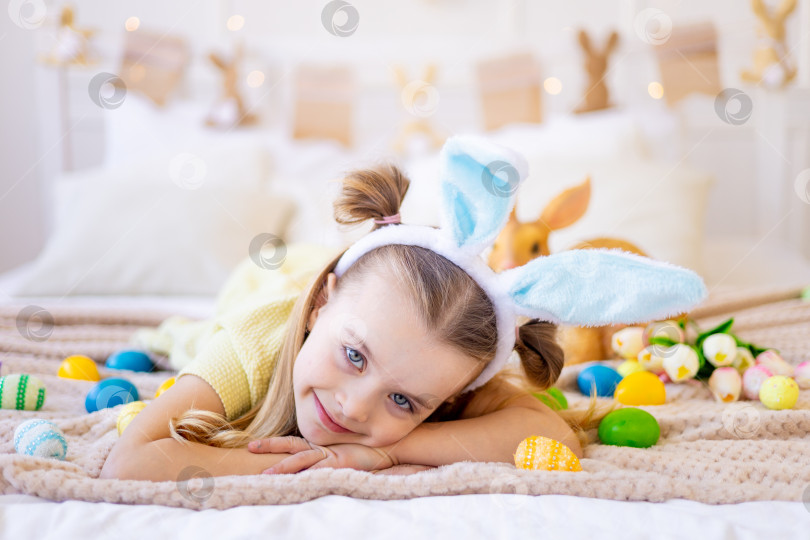 Скачать пасха, маленькая девочка среди раскрашенных яиц в заячьих ушках готовится к празднику, улыбаясь и веселясь фотосток Ozero