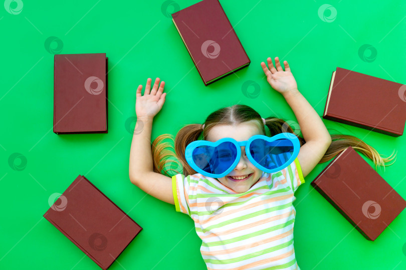 Скачать девочка-ребенок лежит на зеленом фоне с книгами в синих больших забавных очках и улыбается, место для текста фотосток Ozero
