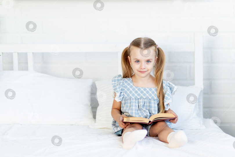 Скачать девочка читает книгу на кровати дома, сидя на белой хлопчатобумажной кровати. фотосток Ozero
