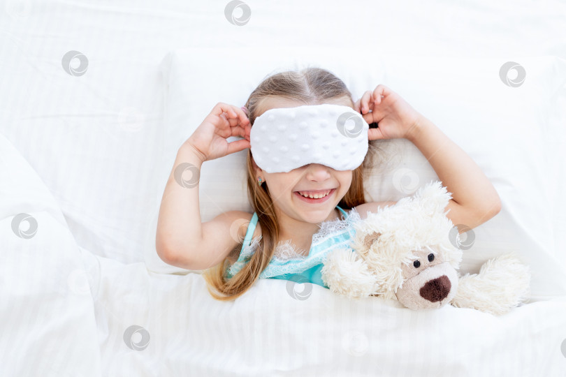 Скачать маленькая девочка в маске для сна проснулась утром дома на кровати, застеленной белым хлопком, с игрушечным плюшевым мишкой и милой улыбкой фотосток Ozero