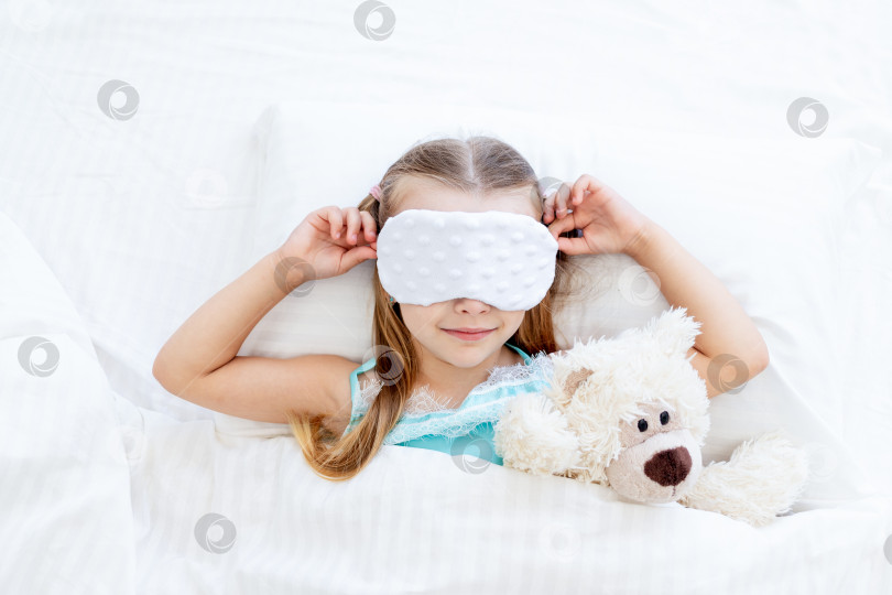 Скачать маленькая девочка в маске для сна проснулась утром дома на кровати, застеленной белым хлопком, с игрушечным плюшевым мишкой и милой улыбкой фотосток Ozero
