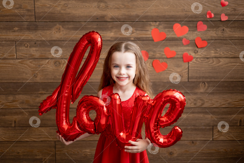 Скачать маленькая девочка в красном платье держит в руках большой воздушный шар с надписью Love на темно-коричневом деревянном фоне с красными сердечками - концепция дня святого Валентина фотосток Ozero