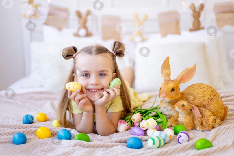 Скачать пасха, маленькая девочка с раскрашенными крашеными яйцами, тюльпанами и кроликом дома в светлой комнате готовится к празднику, улыбается, веселится и играет с яйцами, закрыв глаза фотосток Ozero