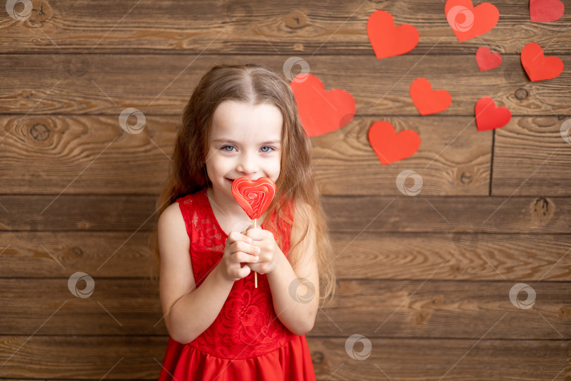 Скачать маленькая девочка в красном платье облизывает языком большой леденец в форме красного сердца на темно-коричневом деревянном фоне с красными сердечками, концепция дня святого Валентина, место для текста фотосток Ozero